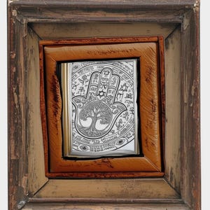 Puede incluir: Una obra de arte enmarcada con una detallada ilustraci&oacute;n en blanco y negro de una mano de Hamsa con un dise&ntilde;o de &aacute;rbol de la vida. La obra est&aacute; dentro de un marco de cuero marr&oacute;n, a su vez dentro de un marco de madera r&uacute;stica m&aacute;s grande.