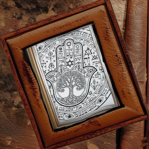 Puede incluir: Una obra de arte enmarcada con un dise&ntilde;o detallado en blanco y negro de la mano de Hamsa con un &aacute;rbol de la vida en el centro. La obra est&aacute; enmarcada en un marco de cuero marr&oacute;n, mostrando un tema espiritual.