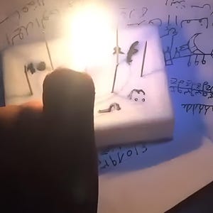Op de afbeelding: Een close-up van een wit, rechthoekig object met zwarte markeringen en lijnen. Het object lijkt klein, mogelijk handgemaakt. De achtergrond bevat handgeschreven tekst en symbolen, wat wijst op een mystiek of artistiek thema.