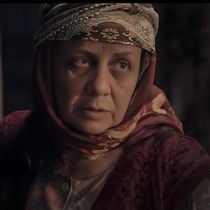Puede incluir: Una mujer con un pañuelo en la cabeza y un chaleco burdeos. El pañuelo tiene diseños florales y el chaleco tiene una apariencia texturizada. La mujer tiene una expresión seria.