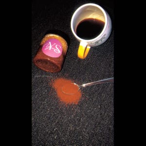 Puede incluir: Un frasco con una etiqueta rosa con las letras "XS" y una taza de café con un asa amarilla están sobre una superficie negra. Una cuchara está al lado de un montón de polvo marrón.