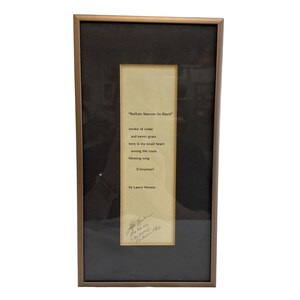 May include: Framed artwork featuring a poem titled "Buffalo Marrow On Black" by Lance Henson. The poem is printed on a cream-colored paper, set against a black background, and framed in a bronze-colored frame. The poem's text is in black.