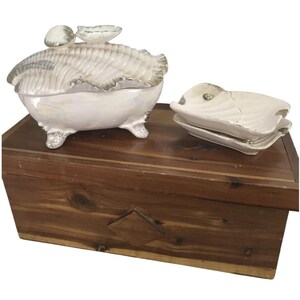 May include: A white ceramic seashell-shaped trinket box with a lid, silver trim, and three feet. Beside it are two matching seashell-shaped dishes. The items are displayed on a wooden chest.