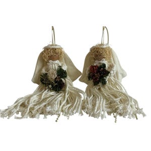 May include: Two handmade angel ornaments. Each angel has a cream-colored fabric body, straw-like hair, and a small bouquet of flowers. The angels have long, fringed skirts and gold hanging loops.
