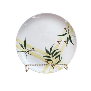 May include: White ceramic plate with a bamboo design. The plate features green bamboo leaves and yellow bamboo stalks. The plate is displayed on a decorative metal stand. The design is painted on the plate.