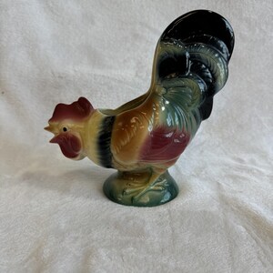 May include: A ceramic rooster figurine in shades of red, yellow, brown, green, and black. The rooster has a detailed comb and tail feathers. The figurine is on a green base and has a small opening on its back.