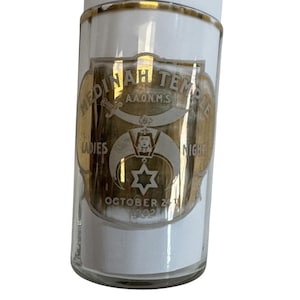 May include: A clear glass container with a white base and a gold-colored emblem. The emblem reads "Medinah Temple A.A.O.N.M.S. Ladies Night October 24 1902". The container has a gold rim.
