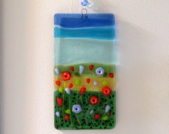 May include: A rectangular glass panel with a layered design depicting a blue sky, green grass, and red and white flowers. The panel is hanging from a white string with a blue and green crystal bead.