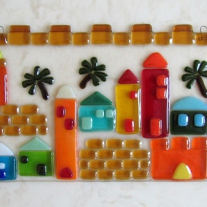 May include: A stained glass wall hanging depicting a cityscape with colorful houses, palm trees, and a brown brick wall. The houses are in shades of red, orange, yellow, blue, and green.
