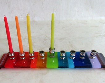 May include: A rainbow-colored glass menorah with nine candle holders and three lit candles. The menorah is made of glass and has a silver finish.