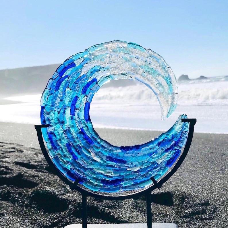 May include: A glass wave sculpture in shades of blue and white, mounted on a black metal stand. The artwork is displayed on a beach with the sea in the background. The sculpture is made of individual glass pieces, creating a textured effect.