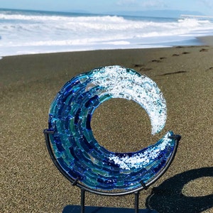 May include: A decorative glass wave sculpture in shades of blue and white, mounted on a black metal stand. The artwork depicts a breaking wave, with the ocean and beach in the background. The sculpture is a decorative piece.