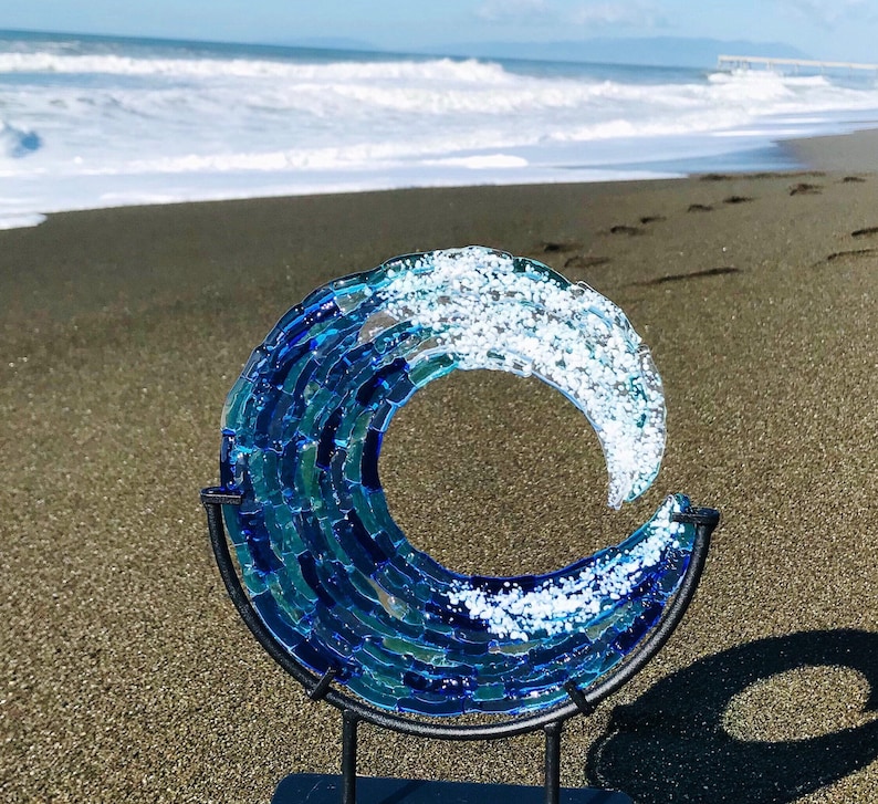 May include: A decorative glass wave sculpture in shades of blue and white, mounted on a black metal stand. The artwork depicts a breaking wave, with the ocean and beach in the background. The sculpture is a decorative piece.