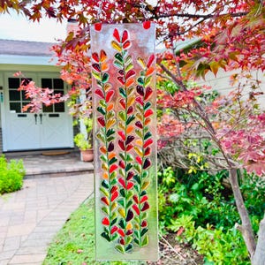 May include: A rectangular glass suncatcher featuring three vertical stems with numerous small, teardrop-shaped glass pieces in shades of red, orange, yellow, and green. The piece is suspended by a silver metal chain attached to two red loops.