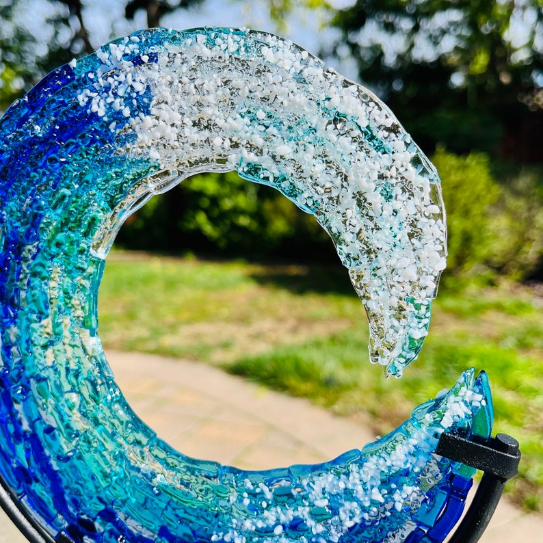 May include: A glass wave sculpture in shades of blue and white. The glass has a textured finish and is mounted on a black metal stand. The colours transition from deep blue to pale blue and white, creating a visual representation of a wave. The sculpture is a decorative item.