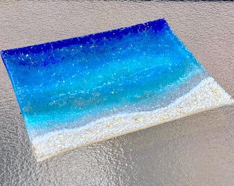May include: A rectangular glass tray featuring a gradient design that transitions from deep royal blue at the top to a lighter turquoise and clear water, ending in a textured white sandy beach edge with small embedded shell fragments.