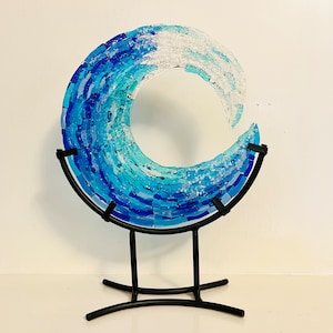 May include: A decorative glass wave sculpture in shades of blue and white, mounted on a black metal stand. The wave is crafted from mosaic-style glass pieces, creating a textured, artistic effect. The stand has a curved design.