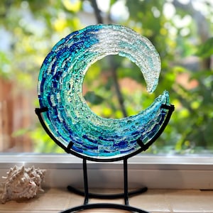 May include: A glass wave sculpture in shades of blue and green, mounted on a black metal stand. The artwork is displayed near a window. The sculpture's dimensions are approximately 20 cm in diameter. A seashell is in the foreground.