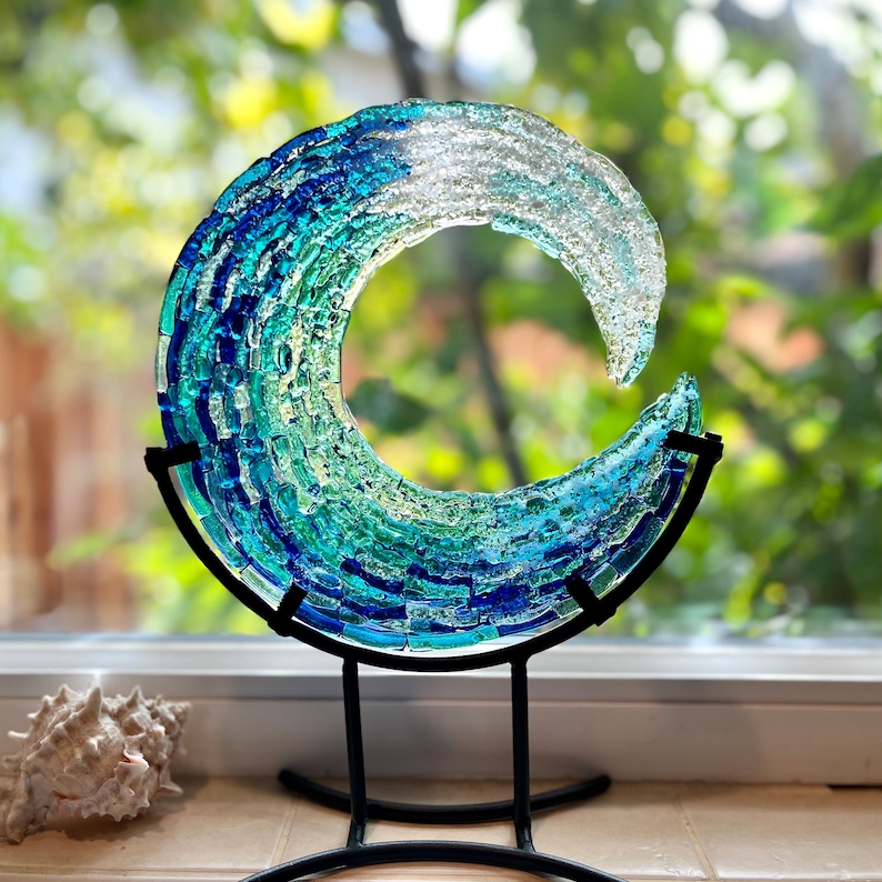 May include: A glass wave sculpture in shades of blue and green, mounted on a black metal stand. The artwork is displayed near a window. The sculpture's dimensions are approximately 20 cm in diameter. A seashell is in the foreground.
