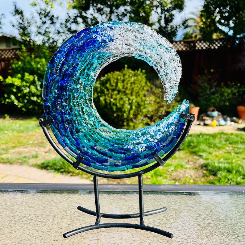 May include: A decorative glass wave sculpture in shades of blue, teal, and white, mounted on a black metal stand. This art piece is designed to resemble a wave. The sculpture is a unique home decor accent, adding a touch of the ocean to any room.