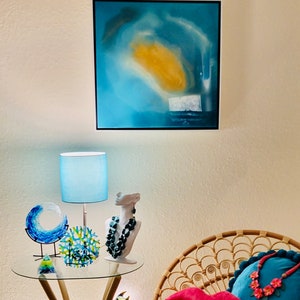 May include: An interior scene with a glass-topped table displaying decorative glass art, a lamp, and a bust with a necklace. A rattan chair holds pillows, including a pink lip-shaped pillow. A painting and blue ribbon decorates the wall.