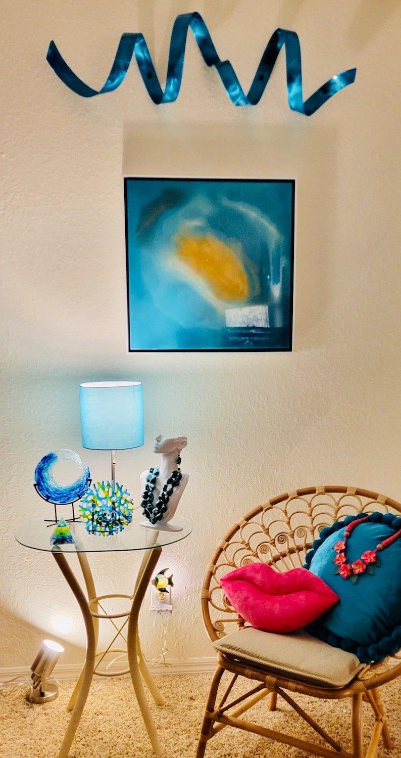May include: An interior scene with a glass-topped table displaying decorative glass art, a lamp, and a bust with a necklace. A rattan chair holds pillows, including a pink lip-shaped pillow. A painting and blue ribbon decorates the wall.