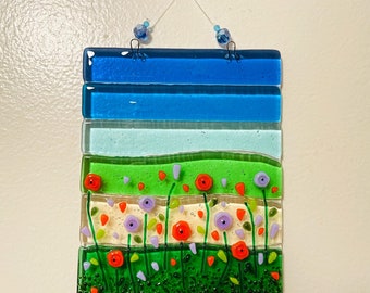 May include: A glass wall hanging with a layered design. The top layer is blue, the next layer is light blue, the next layer is green, and the bottom layer is white with red and purple flowers. The flowers are surrounded by green leaves.