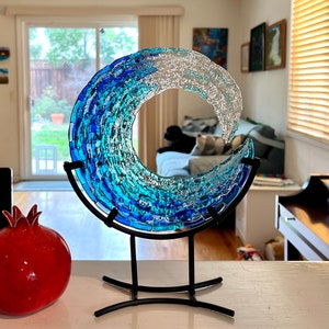 May include: A glass wave sculpture in shades of blue and clear glass, mounted on a black metal stand. The artwork is reminiscent of the ocean. A red pomegranate is in the foreground.