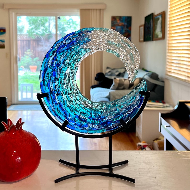 May include: A glass wave sculpture in shades of blue and clear glass, mounted on a black metal stand. The artwork is reminiscent of the ocean. A red pomegranate is in the foreground.