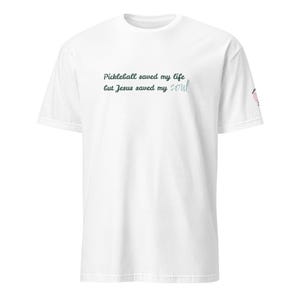 May include: White t-shirt with the text "Pickleball saved my life but Jesus saved my soul" in dark teal and light teal script. The shirt has a crew neck and short sleeves. A small graphic is on the right sleeve.