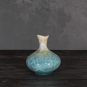 May include: A ceramic vase with a bulbous base and a narrow neck, featuring a crystalline glaze that transitions from a deep teal at the bottom to a lighter blue and soft peach hue near the rim, showcasing intricate star-like crystal formations.