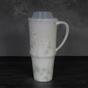 May include: A tall, cylindrical ceramic travel mug with a glossy off-white glaze featuring subtle, light gray circular crystalline patterns. The mug includes a translucent, fitted plastic lid and a curved handle on the right side of the body.