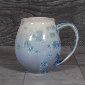 May include: A ceramic mug with a rounded body and a curved handle. The mug features a crystalline glaze that transitions from a creamy white at the top to a soft lavender and blue at the base, decorated with circular blue crystal patterns.