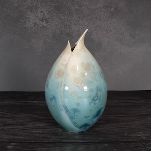 May include: A teardrop-shaped ceramic vase with a glossy finish, featuring a gradient of soft cream at the top transitioning into shades of light and deep blue. The surface displays circular crystalline glaze patterns throughout the body.