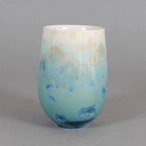 May include: A tall, oval-shaped ceramic tumbler with a unique crystalline glaze. The colors transition from a light beige at the rim to a turquoise and deep teal at the base. The surface features a pattern of circular, crystal-like formations.