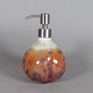 May include: A decorative soap dispenser with a round, textured ceramic body. The body features a blend of orange, brown, and cream colours with a speckled pattern. It has a silver-coloured pump and spout.