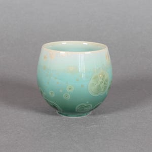 May include: A ceramic cup with a rounded shape and a glossy finish. The cup transitions from a light turquoise at the top to a deeper teal at the base. It features a speckled pattern with light green accents.
