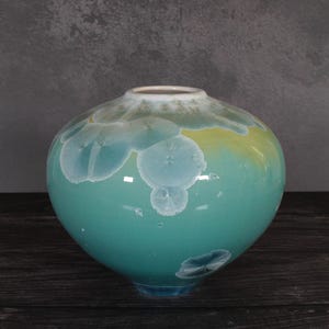May include: A round ceramic vase with a smooth, glossy finish. The body features a vibrant teal base color that transitions into a soft yellow hue near the top. It is decorated with white, crystalline, flower-like patterns scattered across the surface.