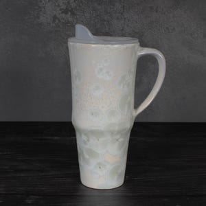 May include: A tall ceramic travel mug with a glossy, off-white glaze featuring a crystalline pattern of soft, circular, pale grey and beige spots. The mug has a sturdy handle on the right side and a translucent, light grey plastic lid with a sip opening.
