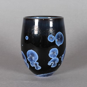 Könnte beinhalten: Ein schwarzer Keramikbecher mit glänzendem Finish und runder Form. Der Becher hat einen dunkelblauen Rand und ist mit hellblauen, organisch geformten Kristallmustern verziert. Die Gesamtästhetik ist modern und künstlerisch.