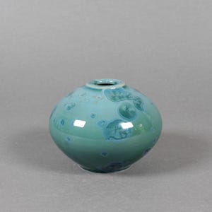 May include: A small, round, teal-colored ceramic vase with a glossy finish. The vase has a narrow opening and is decorated with lighter blue, crystalline-like patterns. The overall shape is spherical, and the vase sits on a solid base.