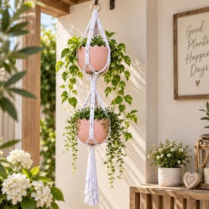May include: A two-tier white macrame plant hanger featuring a wooden ring at the top and a tassel at the bottom. It holds two light pink scalloped pots, each containing a green trailing plant. The hanger is suspended vertically.