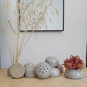 Puede incluir: Una colección de pequeños y redondos ranas de cerámica para flores en un color neutro, algunos con múltiples agujeros para arreglar tallos. Una rana contiene flores rojas secas. La palabra "ongilla" es visible. Sobre una superficie de madera.