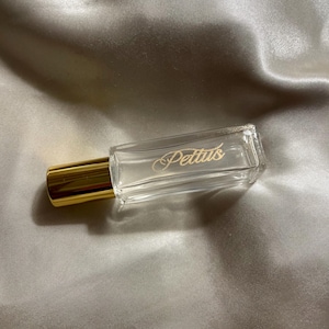 May include: A rectangular clear glass perfume bottle with a gold-colored cap. The front of the bottle features the word Pettus printed in a gold script font. The bottle has a smooth, transparent finish and a metallic gold cylindrical lid.