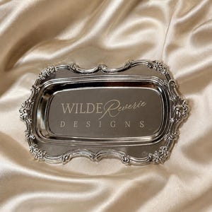 May include: A rectangular silver-toned metal tray with an ornate, scalloped border featuring floral detailing. The center of the tray is smooth and reflective, engraved with the text WILDE Reverie DESIGNS in a combination of script and serif fonts.