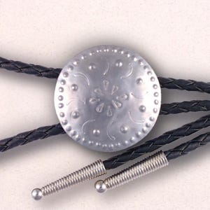 May include: A silver-colored bolo tie with a decorative circular centerpiece. The centerpiece has an embossed floral design and is surrounded by small dots. The black braided leather cord is secured with silver-colored metal tips.