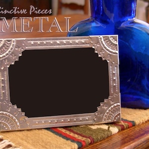 May include: A rectangular silver-colored metal picture frame with embossed geometric patterns and a black center. The frame features decorative corner accents and a dotted border design. The text Distinctive Pieces METAL is visible.