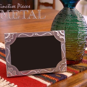May include: A silver metal picture frame with a black interior sits on a colorful woven textile. The frame has decorative corner and edge detailing. A textured green and blue glass vase is in the background. The words "Distinctive Pieces METAL" are at the top.