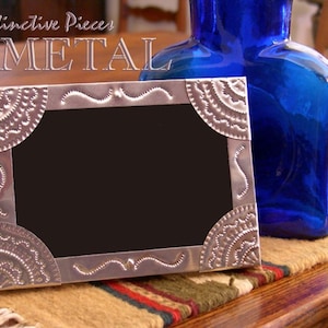 May include: A rectangular metal picture frame featuring embossed corner details with semi-circular patterns and wavy line accents along the edges. The frame has a black center insert and is positioned next to a cobalt blue glass bottle.