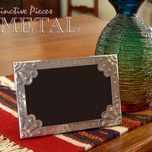 May include: A silver metal picture frame with a black interior, adorned with decorative corner accents. The frame is displayed on a red, white, and black patterned textile, alongside a textured green and blue glass vase. The text "Distinctive Pieces METAL" is visible.